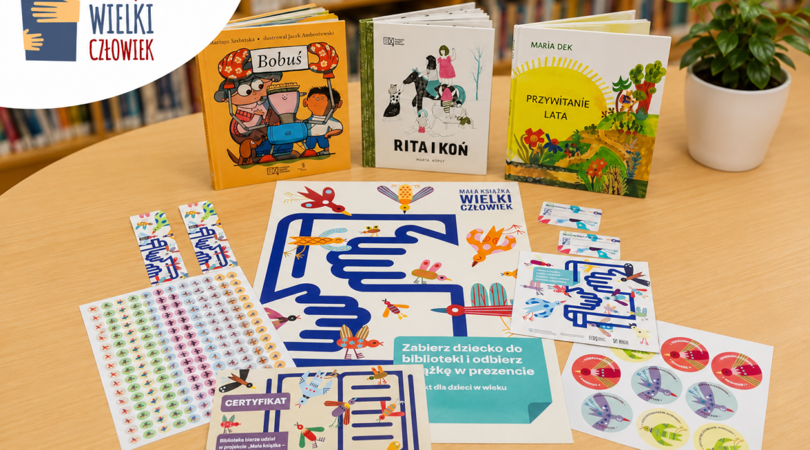Książki dla dzieci, karta małego czytelnika, certyfikat, naklejki i plansze z zadaniami programu „Mała książka – wielki człowiek” ułożone na stole w bibliotece, na tle regałów z książkami.
